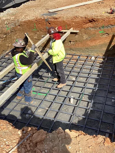Construction workers installing wooden planks over reinforced steel grid in a deep foundation trench at a building site.