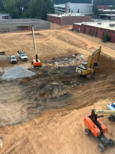 Construction site with heavy machinery including excavators and lifts on cleared earth near industrial buildings.