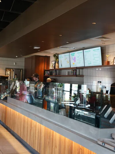 Modern coffee shop interior with wooden counter, digital menu screens, baristas preparing drinks, and a customer in pink outfit