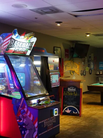 Arcade room with illuminated Baseball Pro and Justice arcade machines next to a pool table in a dim bar.