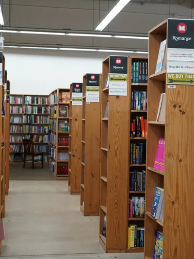 A bookstore aisle with wooden shelves labeled Political Science, Mystery, and Romance filled with books under bright overhead lights.