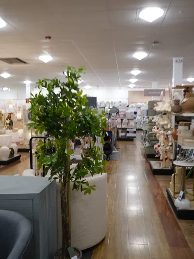Indoor home decor store aisle with artificial tree, furniture, lamps, and bedding items on wooden floor.