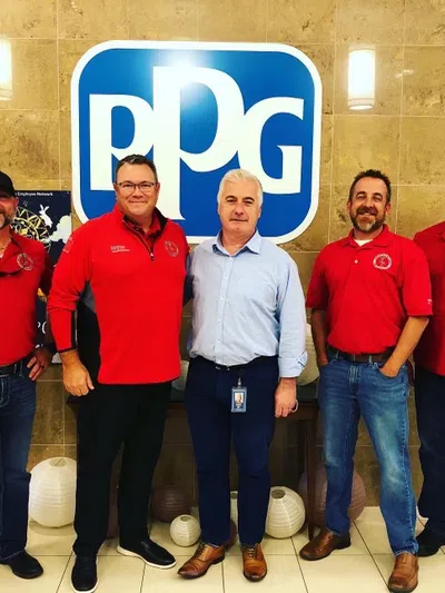 Five men standing in front of a large PPG logo on a beige tiled wall, four in red shirts and one in a blue shirt.