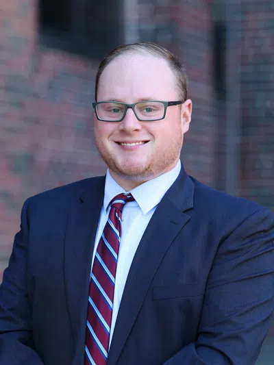 Smiling young man wearing glasses, navy suit, and striped tie posing outdoors with brick wall background.