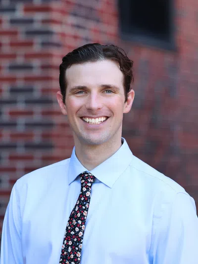 Smiling man wearing light blue shirt and floral tie standing in front of a blurred brick wall background.