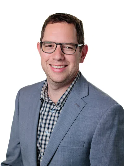 Smiling man wearing glasses, gray blazer, and checkered shirt against a white background.