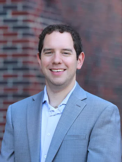 Smiling man in gray suit jacket and light blue shirt stands against a blurred brick wall background.