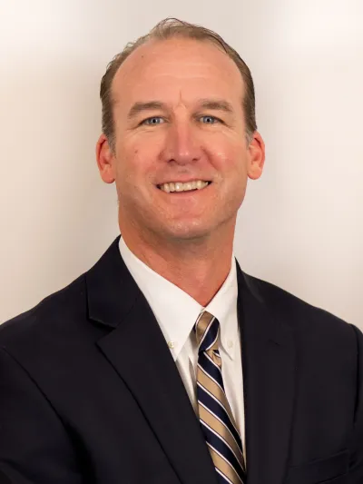 Smiling man in a dark suit jacket, white shirt, and striped tie against a plain light background