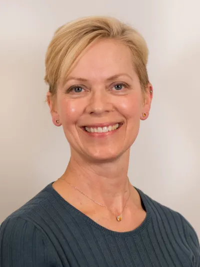 Smiling woman with short blonde hair wearing a dark ribbed sweater and a gold pendant necklace.