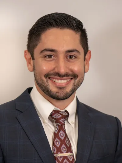 Smiling man with dark hair in a blue suit and patterned red tie against a plain background