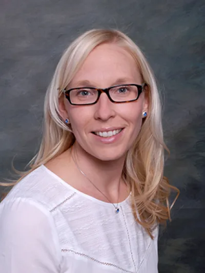 Smiling blonde woman with glasses wearing a white blouse and blue jewelry against a gray backdrop