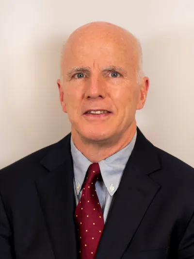 Portrait of a bald man in a dark suit, blue shirt, and red tie with yellow dots against a plain background