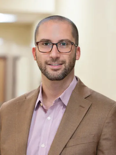 Man with glasses wearing a brown blazer and light purple shirt smiling in a bright indoor setting