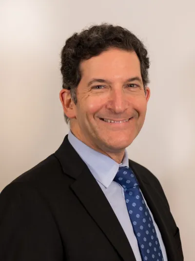 Smiling man with curly hair wearing a dark suit, light blue shirt, and patterned blue tie against a plain background
