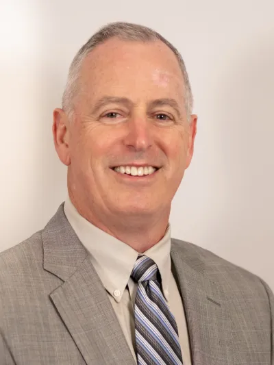 Smiling middle-aged man wearing a gray suit, white shirt, and striped tie on a neutral background
