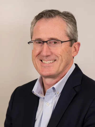 Middle-aged man with glasses smiling, wearing a navy blazer and light blue shirt