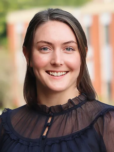 Smiling young woman with blue eyes and dark hair wearing a sheer navy blouse outdoors with blurred background.
