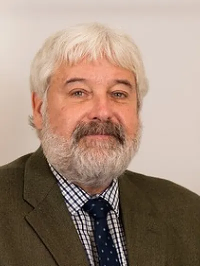 Portrait of a middle-aged man with white hair and beard wearing a checkered shirt, polka dot tie, and brown jacket.