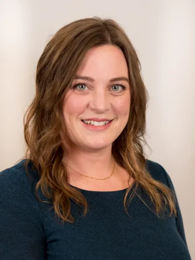 Smiling woman with wavy brown hair wearing a dark teal sweater and delicate gold necklace against a light background