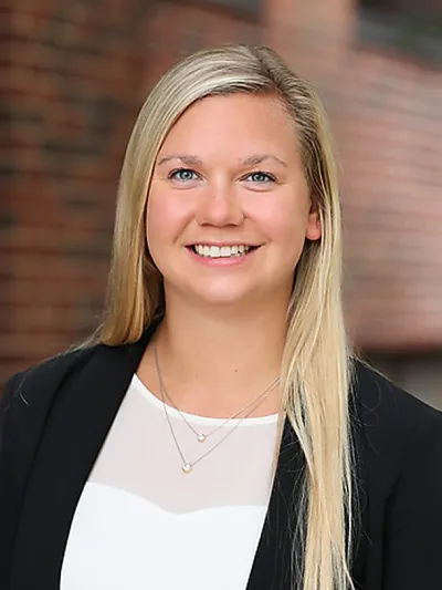 Smiling blonde woman wearing black blazer and white top with layered necklaces against a brick background