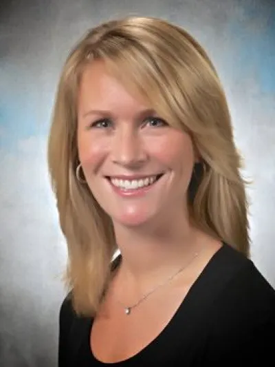 Smiling blonde woman with straight hair wearing a black top and silver necklace against a blue-gray background