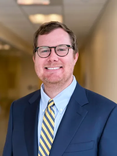 Smiling man wearing glasses, a blue suit, light blue shirt, and yellow striped tie in an indoor setting.