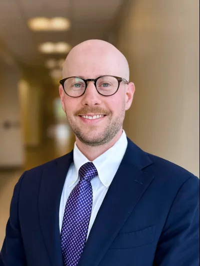 Smiling bald man with glasses wearing a navy suit, white shirt, and purple patterned tie in a softly lit hallway.