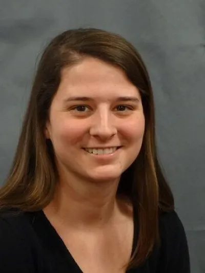 Smiling woman with long brown hair wearing a black top against a gray background.