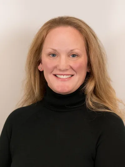 Smiling woman with blonde hair wearing black turtleneck against a plain beige background.
