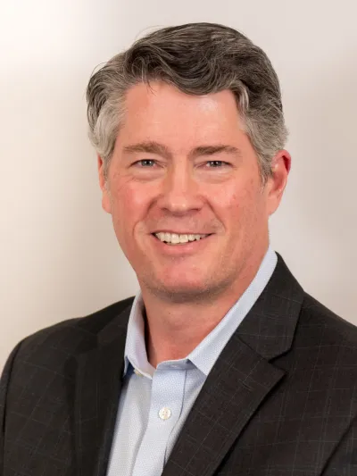 Smiling middle-aged man with gray hair wearing a dark suit jacket and light blue shirt against a plain background
