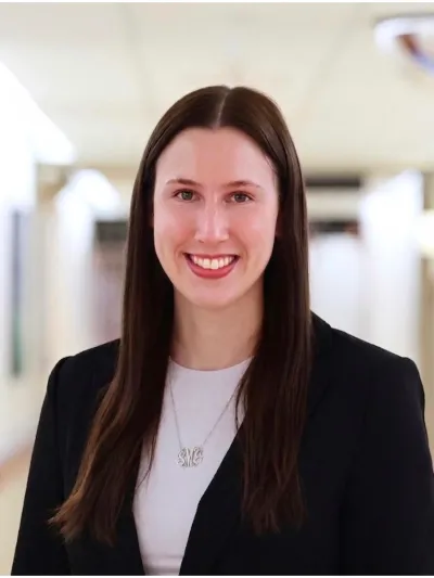 Smiling woman with long brown hair wearing a black blazer and white top in a bright hallway.