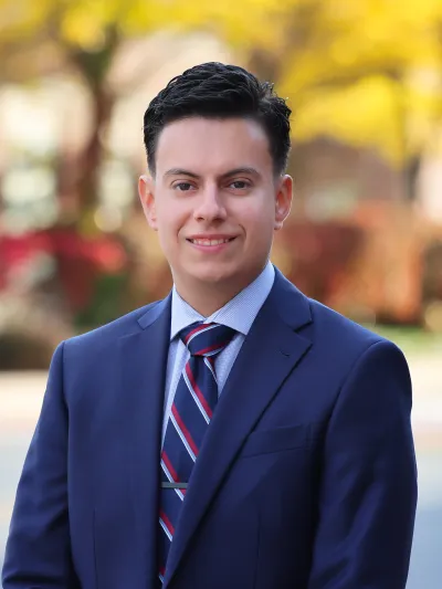 Young man in a navy suit and striped tie smiling outdoors with a blurred colorful background.