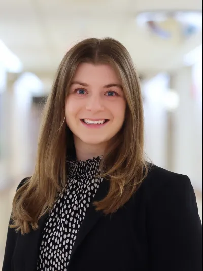 Smiling young woman with long brown hair wearing black blazer and patterned blouse in a bright hallway.