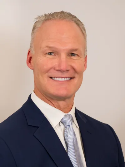 Smiling middle-aged man in a navy suit and light blue striped tie against a plain background.