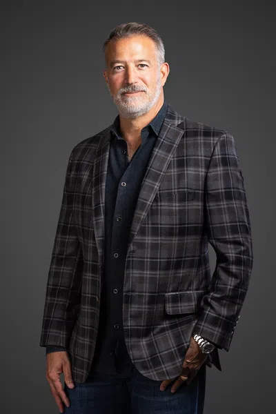 Mature man with grey hair and beard wearing a plaid blazer over dark shirt posing against a gray background