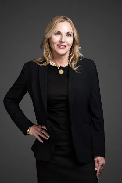 Confident blonde woman in business attire wearing a black dress, blazer, and pearl necklace against a gray background.