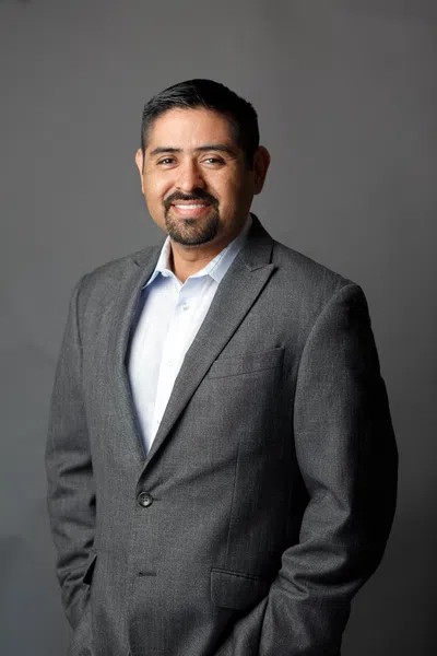 Smiling man in a gray suit jacket and white shirt posing against a neutral gray background.