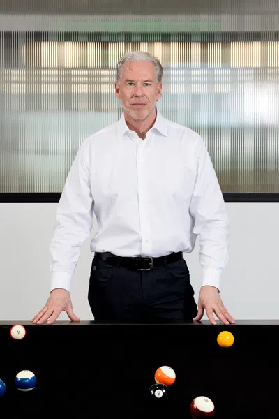 Senior man in white shirt standing behind a black pool table with scattered billiard balls indoors.