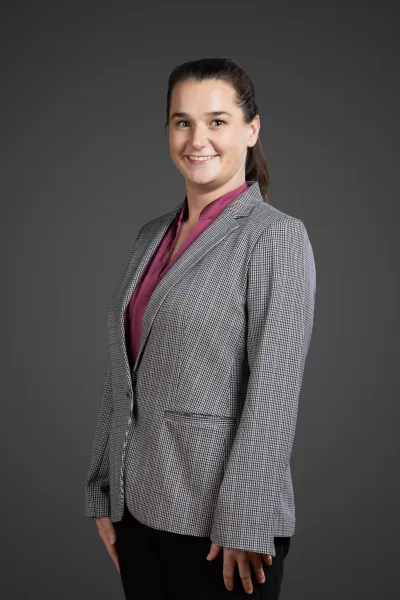 Smiling woman with dark hair in a ponytail wearing a checked blazer and magenta blouse against gray background