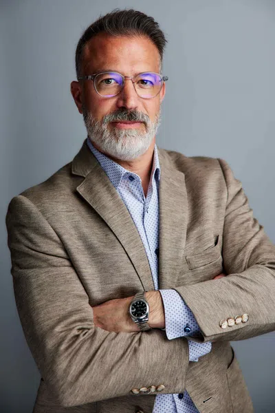 Confident middle-aged man with gray beard and glasses wearing beige blazer and watch, arms crossed.