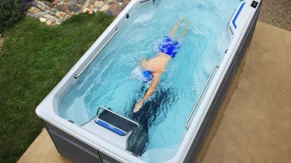 Person swimming in a modern outdoor swim spa with clear water in a garden setting under daylight.