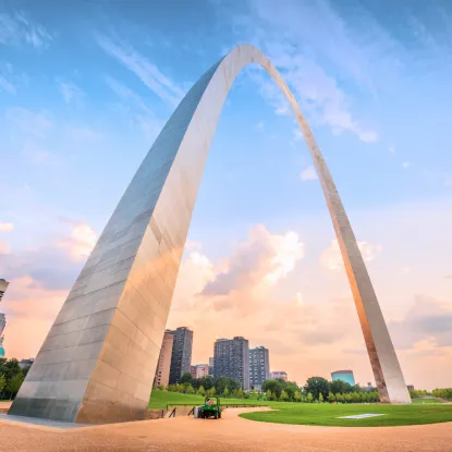 a large arch with grass and trees with Gateway Arch in the background