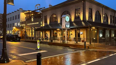 Evening view of a warmly lit brick building with a large clock on the corner and a wet street reflecting lights.