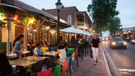Evening view of a lively sidewalk cafe with diners and pedestrians on a brick street under streetlights.