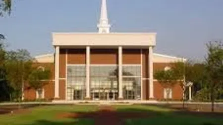 Large red brick church with white columns and tall steeple surrounded by green lawn and trees under clear sky.