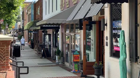 Sunny small town street with shops, storefront awnings, potted plants, and pedestrians walking on sidewalk.