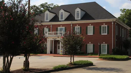Red brick city hall building with white columns, green shutters, and surrounding trees under a blue sky.