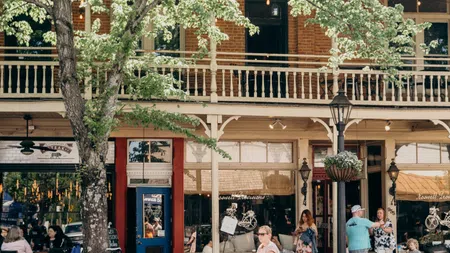 Mother pushing child on toy car while another child rides scooter past bustling café with outdoor seating and trees.