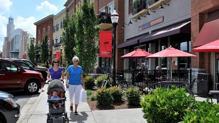 Two women walking with a baby stroller on a sunny urban sidewalk near outdoor café seating with red umbrellas.
