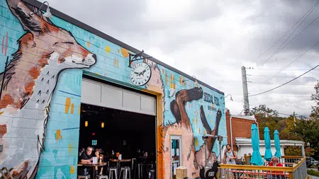 Outdoor brewery with large animal murals on walls and patrons enjoying drinks on a wooden patio under cloudy sky